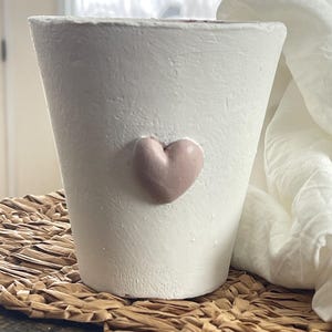 May include: White ceramic planter with a pink heart design. The planter is sitting on a woven brown mat.