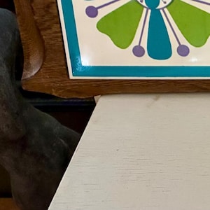 Könnte beinhalten: Ein Vintage-Serviertablett aus Holz mit einem dekorativen Kacheleinsatz. Die Kachel zeigt ein Blumenmuster in Grün-, Blau- und Lilatönen. Das Tablett hat geschnitzte Fächer und eine natürliche Holzoberfläche.