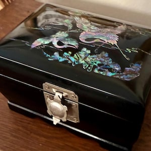 Può includere: Scatola portagioie laccata nera con coperchio incernierato e chiusura argentata. La parte superiore è decorata con intarsi iridescenti in madreperla raffiguranti gru e fogliame. La scatola poggia su una superficie di legno.