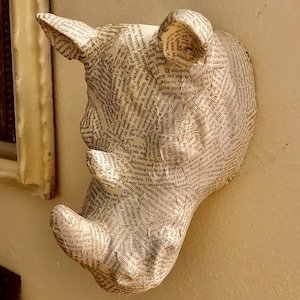 May include: A rhino head sculpture mounted on a wall. The rhino is covered in paper printed with text, giving it a unique, textured appearance. The sculpture is a neutral color, blending with the wall.