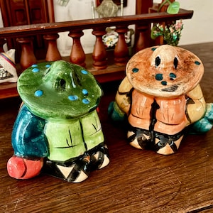 May include: Two ceramic salt and pepper shakers shaped like figures wearing wide-brimmed hats. One figure wears a green hat with blue dots and a green and blue outfit, while the other wears a tan hat with blue dots and a tan and black outfit.