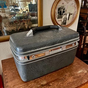 May include: A rectangular, dark gray Samsonite hard-shell train case with a textured finish. The case features a black top handle, silver-toned metal trim, and two integrated key locks on the front panel with the Samsonite brand name clearly visible.