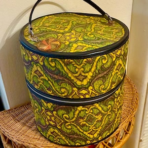 Puede incluir: Sombrerera redonda vintage de dos niveles con asa y ribete negros. La caja presenta un patrón repetitivo en tonos amarillos, verdes y marrones. Un adorno decorativo cuelga del asa. La sombrerera está sobre una mesa auxiliar de mimbre.