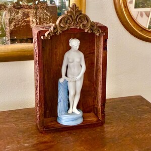 May include: A white porcelain figurine of a standing figure draped in a blue cloth, positioned inside a rectangular wooden frame with a decorative carved top piece. The figure holds a garland of flowers and stands on a circular blue base.