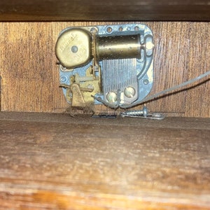 May include: Close-up of the inside of a wooden music box. The music box mechanism is visible, with a metal winding key and a brass gear. The mechanism is attached to a wooden base.