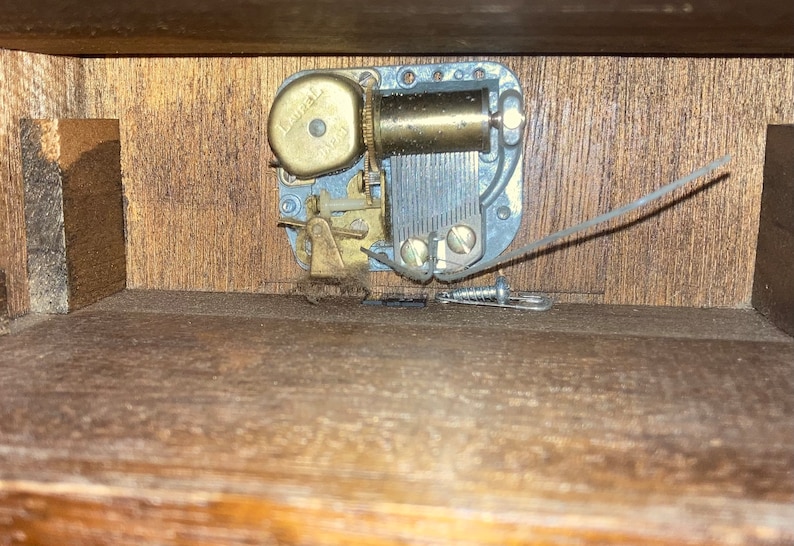 May include: Close-up of the inside of a wooden music box. The music box mechanism is visible, with a metal winding key and a brass gear. The mechanism is attached to a wooden base.