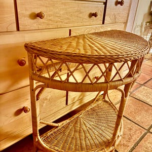 May include: A half-moon shaped wicker side table with a lower shelf. The table is made of light brown woven wicker and has a decorative crisscross pattern on the front. The table is approximately 30 inches wide and 15 inches deep.