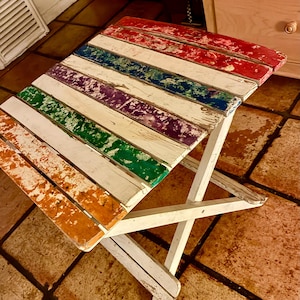 Puede incluir: Pequeña mesa plegable de madera con una superficie multicolor desgastada. La tapa presenta franjas alternas rojas, azules, moradas, verdes, blancas y naranjas. Las patas son blancas y se cruzan para dar soporte. Para uso en exteriores o decoración.