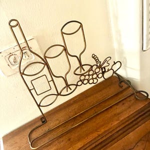 Può includere: Un portabottiglie e bicchieri da vino decorativo in metallo dorato. Il supporto presenta i contorni di una bottiglia di vino, due bicchieri da vino e un grappolo d'uva. Il supporto è progettato per essere posizionato su un tavolo o un bancone.