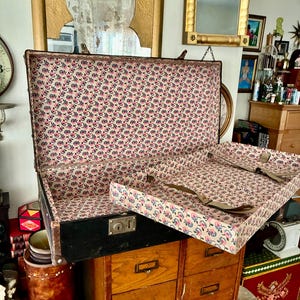 May include: An open antique trunk with a black exterior and a floral patterned interior in pink, white, and blue. The trunk has a removable tray with metal hardware and is set on a wooden cabinet.