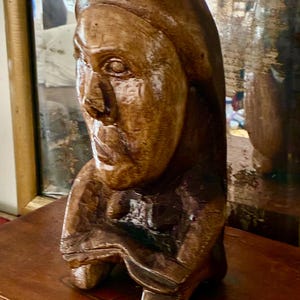 May include: A carved wooden bust of a person with a serene expression. The sculpture is a warm brown color, with visible wood grain and detailed facial features. The bust is set on a wooden surface, with a mirror in the background.