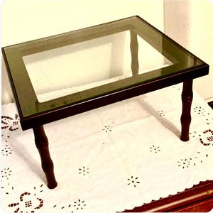 May include: A small, rectangular table with a glass top and dark brown wooden frame. The table has four legs and is approximately 61 cm long, 41 cm wide, and 30 cm high. The table is set on a white tablecloth.