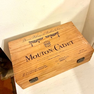 May include: A light brown wooden wine gift box, featuring the text "Mouton-Cadet" and "Baron Philippe de Rothschild". The box has metal clasps and text indicating it is for two bottles of wine, with a Bordeaux origin.