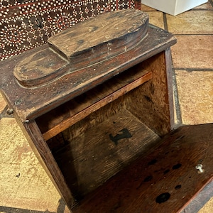 以下が含まれることがあります： 上部に靴型が組み込まれた、アンティークの木製靴磨き箱。箱は濃い茶色で、木目がはっきりと見え、蝶番付きのドアが付いています。内部には小さな棚があります。靴のお手入れや収納に使用される可能性があります。