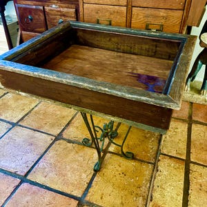 Puede incluir: Un expositor de madera vintage con una bandeja rectangular marrón oscuro y una base decorativa de metal verde. El expositor está colocado sobre un suelo embaldosado. La bandeja tiene un aspecto envejecido, lo que sugiere edad y uso.