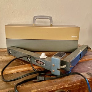 Puede incluir: Un foco de cineasta Smith-Victor vintage y una caja metálica de dos tonos con asa. La luz es gris con dos bombillas y un cable de alimentación negro. La caja es dorada y gris.