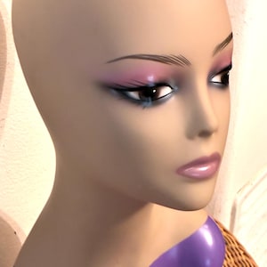 May include: A mannequin head with painted makeup. The mannequin has a neutral skin tone, with detailed eye makeup in shades of pink and blue. The lips are a soft pink. The neck is attached to a purple base.