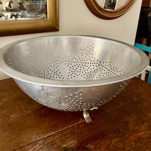 May include: A round, silver-colored metal colander featuring a wide rim and numerous small, evenly spaced drainage holes throughout the bowl. The colander is supported by three small, curved metal feet attached to the base of the exterior.