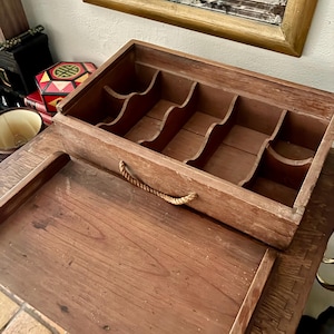 May include: A vintage wooden jewelry box with a tray and multiple compartments. The box is open, revealing the interior with curved dividers. A rope handle is attached to the top. The box is made of dark wood and sits on a wooden table.