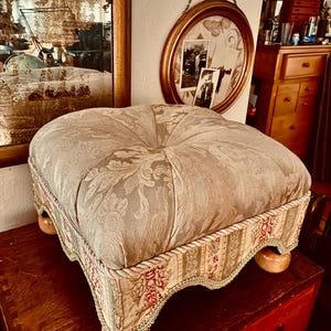 May include: A square tufted ottoman featuring a light beige damask fabric top with a central button. The sides are upholstered in a coordinating striped fabric with red floral accents, finished with decorative rope trim and light wood bun feet.