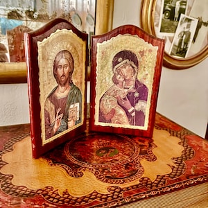 Pode incluir: Dois ícones religiosos com detalhes em folha de ouro. Os ícones estão emoldurados em molduras de madeira vermelha escura. Um retrata uma figura com vestes verdes e marrons, segurando um livro. O outro mostra uma mulher com vestes roxas segurando uma criança. Os ícones são exibidos em um livro decorativo.