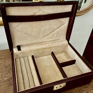 May include: A dark brown jewelry box with a hinged lid and a gold-toned metal latch. The interior is lined with cream-colored fabric, featuring a ring roll section, two open compartments, and a horizontal strap on the inside of the lid.