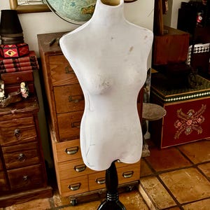 May include: A white dress form with a black stand. The form has a bust and waist shape, and the stand has a tripod base. The dress form is in front of a wooden dresser and a globe.