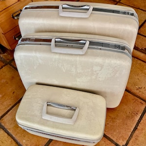 May include: A set of three off-white hard-shell Samsonite suitcases of varying sizes, each featuring a rectangular shape, a top-mounted handle with metallic accents, and a secure locking mechanism along the silver-toned frame of the luggage.
