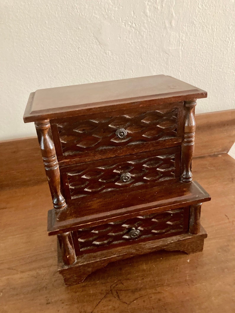 May include: A small wooden dresser with three drawers. The drawers have a carved design and metal knobs. The dresser is a dark brown color.