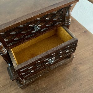 May include: A small wooden jewelry box with two drawers. The drawers have a carved design and are lined with a brown fabric. The box is made of dark wood and has a rustic look.