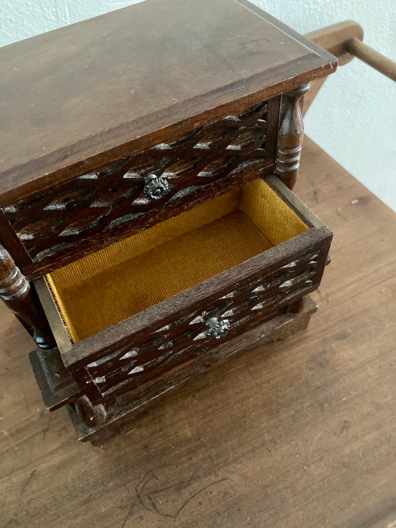 May include: A small wooden jewelry box with two drawers. The drawers have a carved design and are lined with a brown fabric. The box is made of dark wood and has a rustic look.