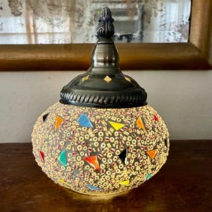 May include: A decorative, round lamp with a mosaic glass shade. The shade features colorful triangular and round glass pieces. The lamp has a dark metal base and a decorative finial on top. The lamp is lit, emitting a warm glow.