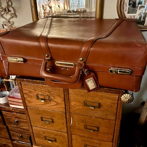 May include: A rectangular brown leather suitcase featuring gold-toned metal latches, a sturdy top handle, and a matching leather luggage tag. The suitcase has a smooth finish with stitched detailing along the front and reinforced corner accents.