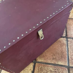 May include: A burgundy-colored storage trunk with a rectangular shape. The trunk is adorned with small, silver-colored rivets along the edges and features a brass-colored clasp. The trunk appears to be made of a leather-like material.