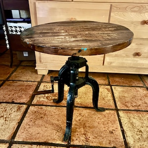 May include: A round, wooden-topped side table with a distressed finish. The table has a black, industrial-style base with three curved legs and a hand crank for height adjustment. The tabletop is approximately 18 inches in diameter.