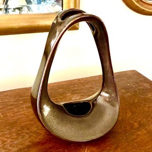 May include: A dark brown ceramic vase with a smooth, glossy finish and a distinctive open, arched handle design. The base is wide and curved, tapering upward into a singular, continuous loop that forms the handle and the central opening.