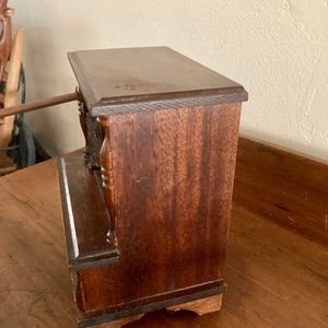 May include: A small wooden box with a dark brown finish. The box has a hinged lid and a decorative front panel. The box is sitting on a wooden surface.