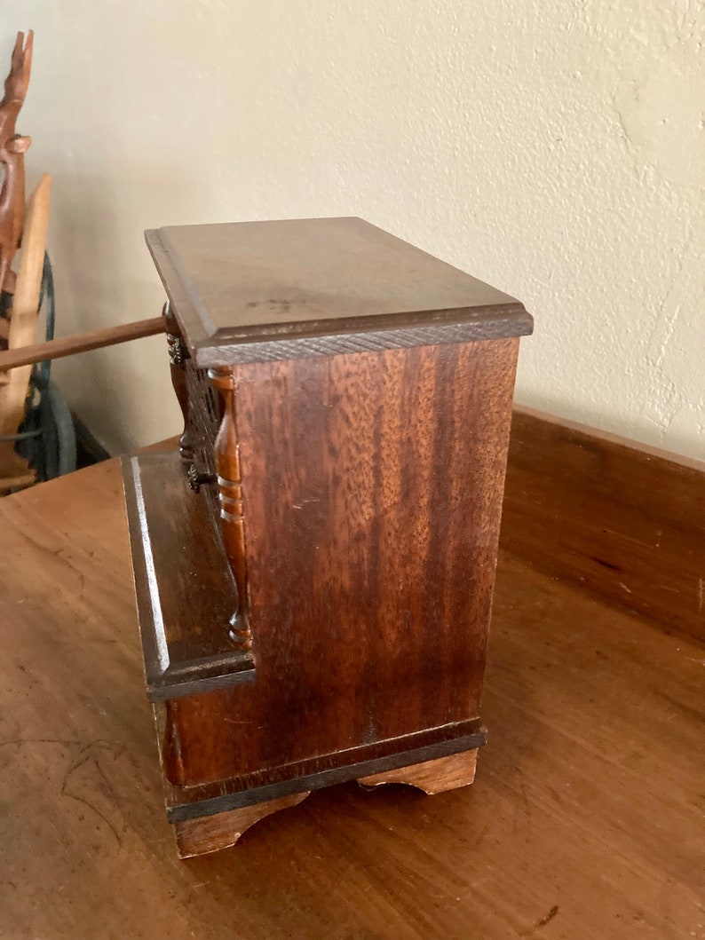 May include: A small wooden box with a dark brown finish. The box has a hinged lid and a decorative front panel. The box is sitting on a wooden surface.
