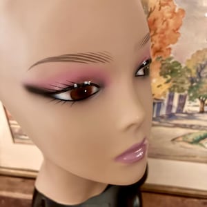 May include: A mannequin head with detailed makeup. The eyes feature dark eyeliner, pink eyeshadow, and small rhinestones. The lips are a glossy, light purple. The head is on a black base, with a neutral skin tone.
