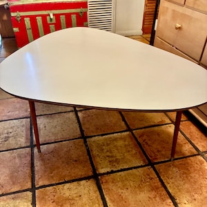 May include: A mid-century modern coffee table with a white, asymmetrical top and four reddish-brown legs. The table has a dark brown trim around the edge of the tabletop. The legs are angled and taper towards the floor. The table is approximately 40 inches long.