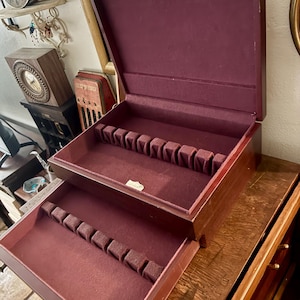 May include: A vintage wooden silverware chest with a burgundy velvet interior. The chest has two levels with slots for utensils. The open top reveals the interior, showcasing the storage design. The exterior is a rich, dark wood.