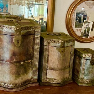 May include: A set of three graduated storage boxes with a weathered, multi-toned finish in shades of green, brown, and gray. Each box features a hinged lid, a metal handle on top, a heart-shaped latch, and decorative textured metal trim.