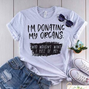 May include: A white t-shirt with the text "I'm donating my organs. Who wouldn't want a piece of me?" printed in black. The shirt is laid out on a wooden surface with a pair of denim shorts, sunglasses, and white sneakers.