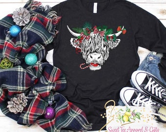 May include: Black long-sleeve t-shirt with a white Highland cow wearing a Christmas wreath and holding a candy cane. The cow has a red nose and is looking at the viewer.