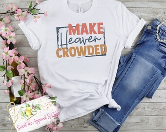 May include: White t-shirt with a blue and orange graphic that says "Make Heaven Crowded". The shirt is folded and laid on a wooden surface with a pair of blue jeans and a bottle of wine.