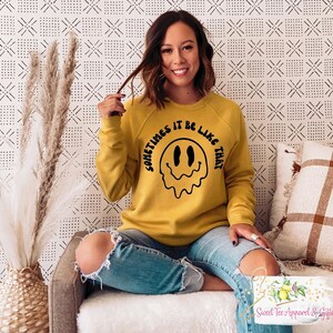 Op de afbeelding: Een geel sweatshirt met een zwarte afbeelding van een smeltende smiley en de tekst "Sometimes it be like that".