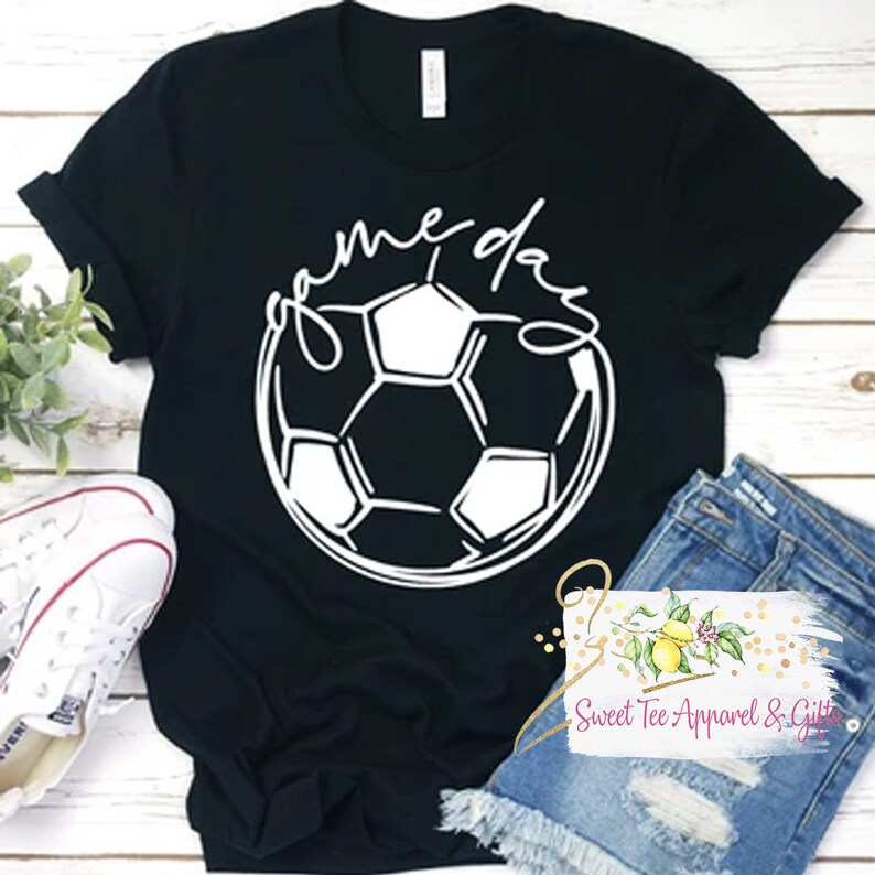 以下が含まれることがあります： 白いサッカーボールのグラフィックと「game day」というテキストが書かれた黒いTシャツ。テキストは筆記体で書かれています。