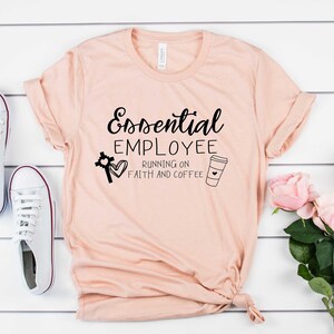 May include: A peach-colored t-shirt with the text "Essential Employee Running on Faith and Coffee" in black. The text is accompanied by a cross with a heart and a coffee cup with a heart.