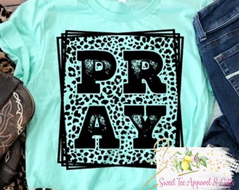 May include: A turquoise t-shirt with a black graphic of the word "PRAY" in a leopard print pattern. The graphic is framed by a black square.