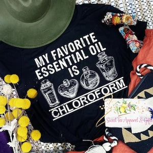 May include: Black t-shirt with white text that reads "My Favorite Essential Oil Is Chloroform" and a graphic of three bottles with a heart shape in the middle.